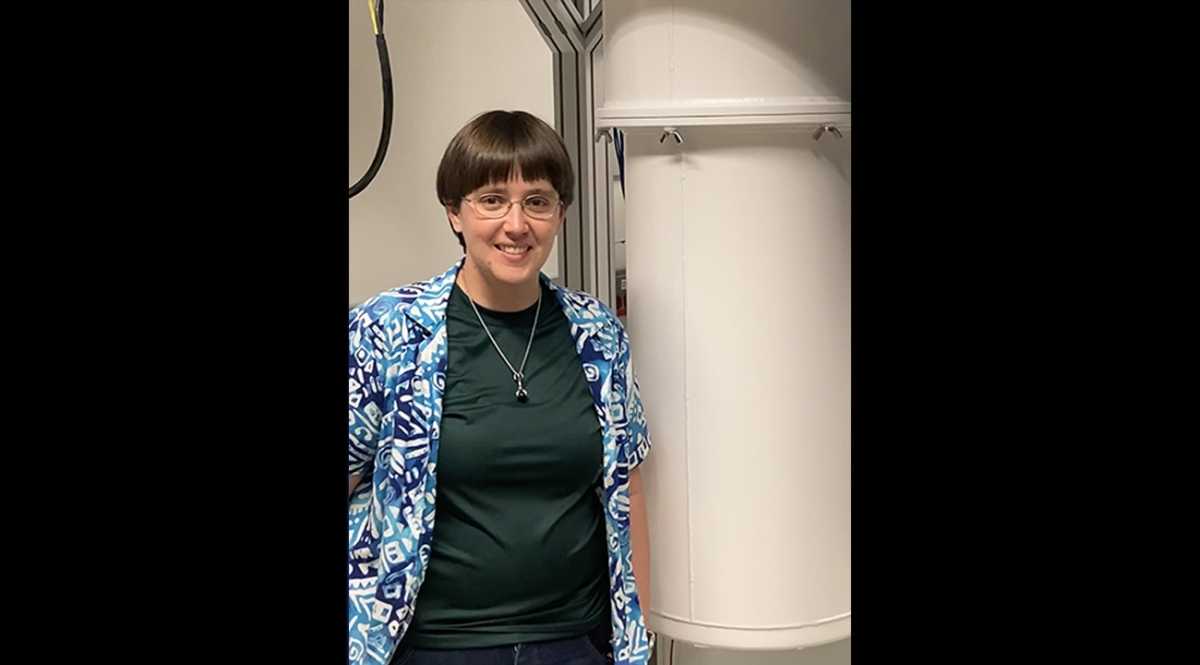 Physicist Alicia Kollar stands next to a white dilution refrigerator that looks like a large, vertical, white pipe.